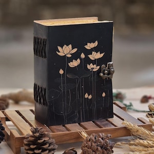 May include: A black journal with a floral lotus design and a metal clasp. The journal has a textured cover and a woven spine. The pages are visible at the top. The journal is sitting on a wooden tray.