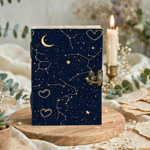 May include: A dark blue journal featuring a gold celestial pattern of stars, constellations, a crescent moon, and heart outlines. The rectangular book includes a metallic bronze-colored latch closure on the right side to keep it securely shut.