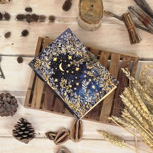 Puede incluir: Un diario decorativo con un dise&ntilde;o celestial. La portada presenta un fondo azul oscuro con una luna creciente, estrellas y detalles florales y de hojas doradas. El diario est&aacute; asegurado con un cierre de metal y descansa sobre una bandeja de madera.