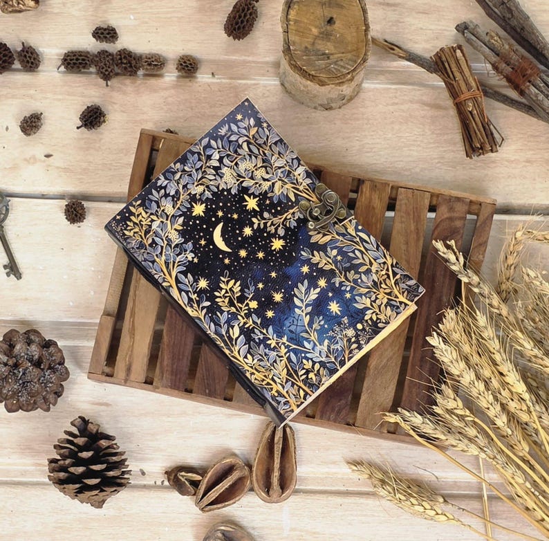 Puede incluir: Un diario decorativo con un dise&ntilde;o celestial. La portada presenta un fondo azul oscuro con una luna creciente, estrellas y detalles florales y de hojas doradas. El diario est&aacute; asegurado con un cierre de metal y descansa sobre una bandeja de madera.