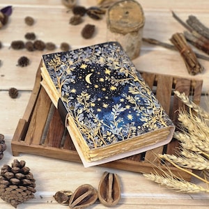 Puede incluir: Un diario hecho a mano con un dise&ntilde;o celestial. La portada presenta un fondo azul oscuro con una luna creciente, estrellas y follaje dorado. El diario est&aacute; encuadernado con una correa de cuero negro y descansa sobre una bandeja de madera.