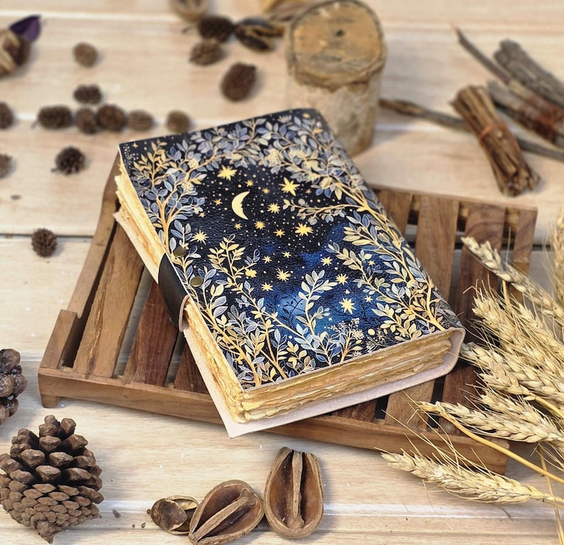 Puede incluir: Un diario hecho a mano con un dise&ntilde;o celestial. La portada presenta un fondo azul oscuro con una luna creciente, estrellas y follaje dorado. El diario est&aacute; encuadernado con una correa de cuero negro y descansa sobre una bandeja de madera.