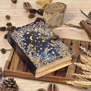 Puede incluir: Un diario azul oscuro con un dise&ntilde;o celestial que presenta una luna creciente y estrellas, enmarcado por ilustraciones bot&aacute;nicas doradas. El diario est&aacute; asegurado con un cierre de metal y descansa sobre una bandeja de madera, rodeado de elementos decorativos naturales.
