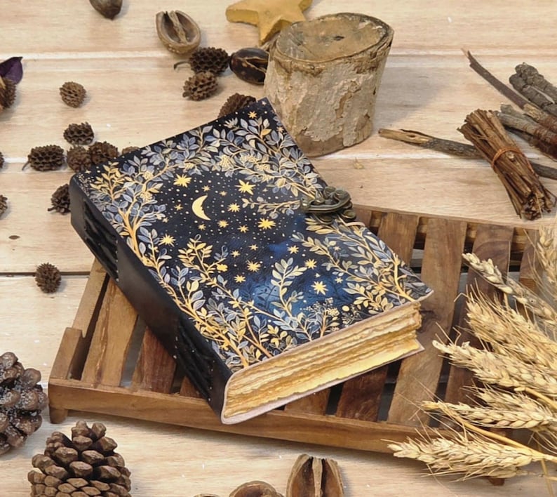 Puede incluir: Un diario azul oscuro con un dise&ntilde;o celestial que presenta una luna creciente y estrellas, enmarcado por ilustraciones bot&aacute;nicas doradas. El diario est&aacute; asegurado con un cierre de metal y descansa sobre una bandeja de madera, rodeado de elementos decorativos naturales.