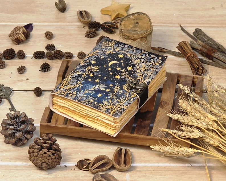 Puede incluir: Un diario de estilo vintage con una cubierta de dise&ntilde;o celestial que presenta una luna creciente y estrellas. El diario abierto revela p&aacute;ginas de papel envejecido. Descansa sobre una bandeja de madera, rodeado de pi&ntilde;as y trigo seco.