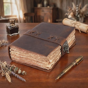 May include: A dark brown leather journal with thick, deckled-edge paper pages. The cover features a wide leather strap secured with a metal latch and decorative cross-stitching. A glass ink bottle and a metal fountain pen rest nearby.