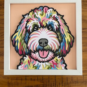 May include: A framed paper art portrait of a dog, featuring a vibrant mix of colours in its fur: pink, yellow, blue, and green. The dog has a black nose and a pink tongue. The background is a soft peach colour, all within a white frame.