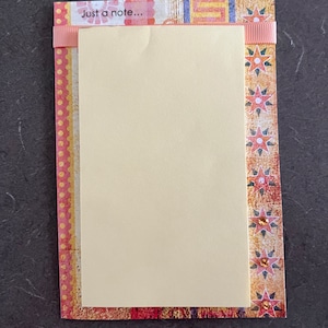 Puede incluir: Una tarjeta en blanco con una cinta rosa y un borde decorativo. La tarjeta tiene el texto "Just a note..." impreso en la parte superior.