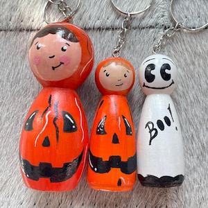 Op de afbeelding: Drie houten poppetje-sleutelhangers. Twee poppetjes zijn beschilderd als oranje pompoenen met zwarte jack-o'-lantern gezichten en bruin haar. Het derde poppetje is wit met een zwart spookgezicht en de tekst Boo! in zwarte letters op de voorzijde.