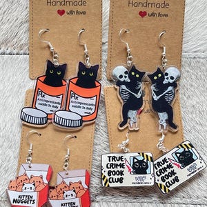 Op de afbeelding: Vier paar acryl bungelende oorbellen op bruine kartonnen kaartjes. De ontwerpen bevatten zwarte katten in oranje medicijnflesjes met het label 'Antidepressants cuddle 3x daily', zwarte katten die skeletten knuffelen, kitten nuggets en 'True Crime Book Club' bordjes.