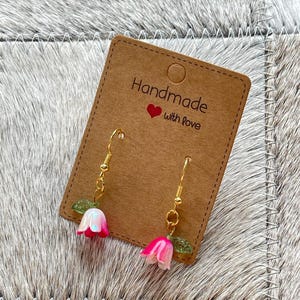 Op de afbeelding: Een paar oorbellen met roze en witte klokvormige bloemhangers en kleine groene blaadjes, hangend aan goudkleurige metalen haakjes. De oorbellen zijn bevestigd aan een bruine rechthoekige displaykaart met de tekst Handmade with love.