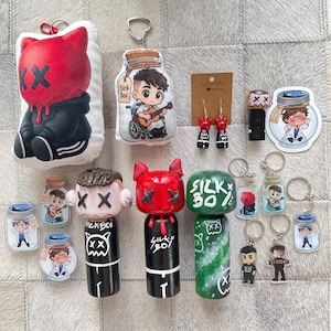 Op de afbeelding: Een verzameling handgemaakte sleutelhangers, stickers en knuffels. De items bevatten stripfiguren met rode en zwarte kleurenschema's, sommige met de tekst "Sick Boi" en "Silky Boy". De items zijn gerangschikt op een getextureerd oppervlak.