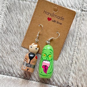 Op de afbeelding: Een paar bungelende oorbellen met handbeschilderde houten poppetjes. De ene hanger toont een figuur in een beige outfit met bruin haar, de andere is een groen, wijdmondig wezen met gele ogen en een roze tong, bevestigd aan zilverkleurige haken.