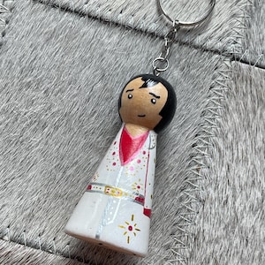 Op de afbeelding: Een houten poppetje sleutelhanger met een handgeschilderde figuur met zwart haar, een rode V-hals en een witte jumpsuit versierd met gouden en rode stervormige accenten, een zilveren riem en een metalen ring bevestigd aan een zilveren ketting.