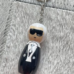 Op de afbeelding: Een houten poppetje sleutelhanger met wit haar, een zwarte zonnebril en een zwart pak met een wit overhemd, zwarte das en een klein wit bloemdetail. Het figuurtje is bevestigd aan een zilverkleurige metalen ketting en een ronde sleutelring.