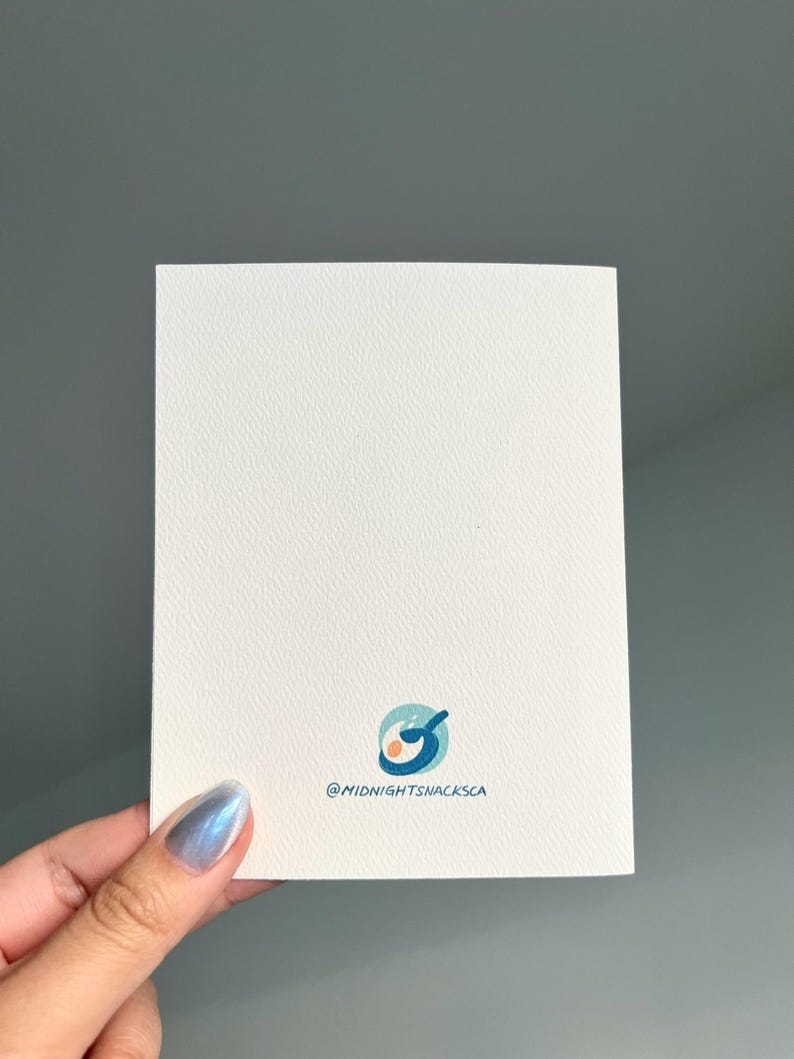 Pu&ograve; includere: Un biglietto bianco con un'illustrazione blu e arancione di una padella con due uova. Il testo "@midnightsnacksca" &egrave; stampato sotto l'illustrazione.