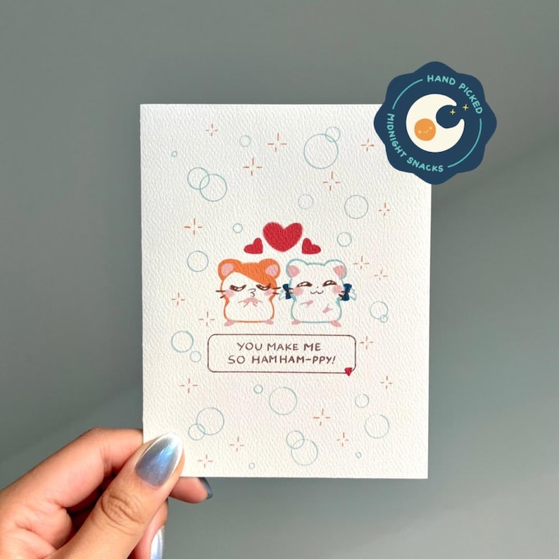 Pu&ograve; includere: Un biglietto bianco con due criceti dei cartoni animati che si tengono per mano e cuori sopra di loro. Il biglietto dice "Mi fai cos&igrave; hamham-ppy!"