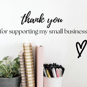 May include: A white wall with the text "thank you for supporting my small business" and a hand-drawn heart. There are three notebooks with gold spiral bindings and a pink notebook on a shelf. A potted plant and a cup of pens are also on the shelf.