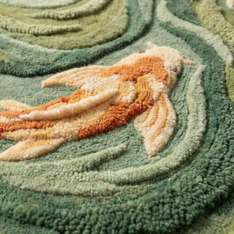May include: A hand-tufted rug featuring a koi fish design. The fish is rendered in shades of orange, cream, and tan, set against a backdrop of green and olive-green waves. The rug's texture is visible, with looped yarn creating a three-dimensional effect.