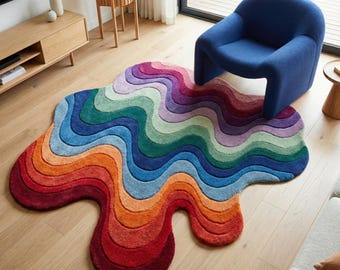 May include: A vibrant, multi-coloured rug with a flowing, organic shape. The rug features a rainbow gradient, transitioning from red to purple. It's placed on a wooden floor, with a blue armchair nearby. The rug's dimensions are approximately 180cm x 120cm.