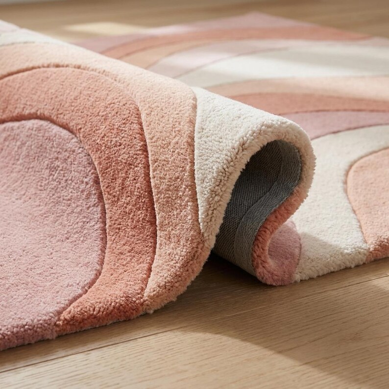 Puede incluir: Una alfombra enrollada con un dise&ntilde;o de arco&iacute;ris en tonos rosa, melocot&oacute;n y crema. La textura de la alfombra es visible, con un pelo suave y rizado. El borde enrollado revela el reverso. La alfombra est&aacute; sobre un suelo de madera.