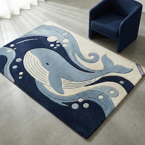 May include: A rectangular rug featuring a whale design in shades of blue and off-white. The whale is swimming in stylized waves. The rug is handmade and has a soft, textured appearance. A blue armchair is in the background.