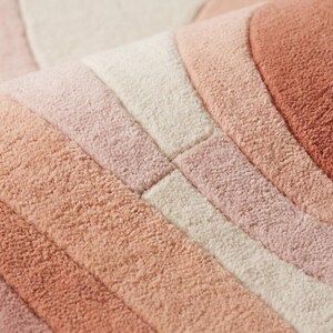 Puede incluir: Primer plano de una alfombra suave y texturizada con un dise&ntilde;o de arco&iacute;ris. La alfombra presenta rayas de diferentes tonos de rosa, melocot&oacute;n y crema. El material afelpado es visible, lo que sugiere comodidad y calidez. El dise&ntilde;o es moderno y acogedor.