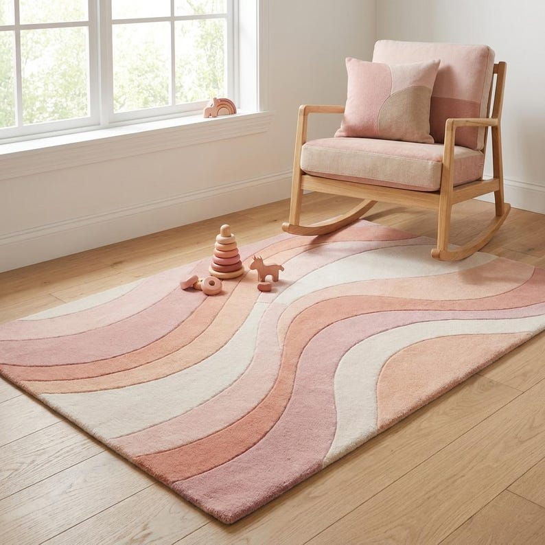 Puede incluir: Una alfombra en tonos rosados con un dise&ntilde;o ondulado en tonos rosa, melocot&oacute;n y crema. Una mecedora de madera con un coj&iacute;n y una almohada rosas est&aacute; cerca. Juguetes de madera, incluyendo una torre apilable y un peque&ntilde;o caballo, est&aacute;n en el suelo.