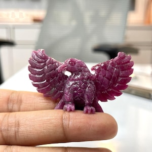 Puede incluir: Una pequeña figura de águila tallada a mano en una piedra preciosa de color magenta intenso. El ave se muestra con las alas completamente extendidas, exhibiendo texturas de plumas detalladas en el cuerpo, un pico afilado y garras definidas.