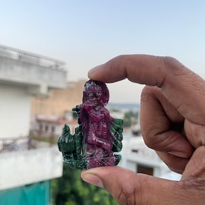 Könnte beinhalten: Eine kleine, geschnitzte Figur einer hinduistischen Gottheit, möglicherweise Krishna, aus einem grünen und rosafarbenen Edelstein. Die Figur hält eine Flöte und steht auf einem Sockel mit einem Pfauenmuster.