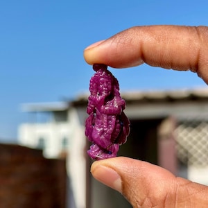 Könnte beinhalten: Eine kleine, aufwendig geschnitzte Figur in tiefem Magenta. Die Figur scheint eine Gottheit oder eine spirituelle Figur mit detaillierten Gesichtszügen und aufwendiger Kleidung darzustellen. Die Schnitzerei wird zwischen Fingern vor einem hellblauen Himmel gehalten.