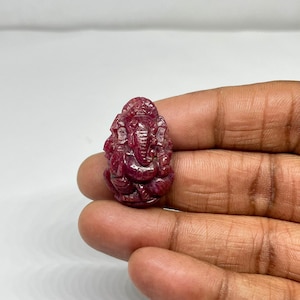 May include: A carved, deep red gemstone figurine of the Hindu deity Ganesha. The figurine is highly detailed, with visible features including the elephant head, multiple arms, and seated posture. The gemstone has a polished, reflective surface.