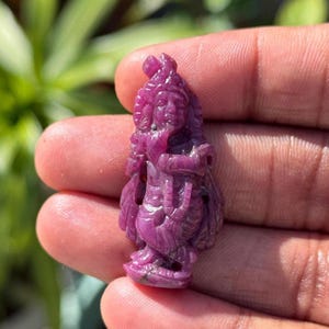 May include: A carved, magenta-colored gemstone figurine of a deity, held in a hand. The figure is detailed with a headdress and ornate clothing. The background is blurred, with green foliage.