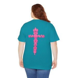 May include: A teal t-shirt featuring a pink cross design on the back. The cross is intricately detailed with various seashell shapes. The shirt is made of a soft material and has a classic crew neck.