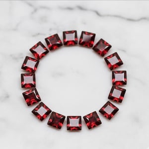 May include: A collection of square-cut, deep red gemstones arranged in a circular pattern. The gems are set against a white marble background, showcasing their vibrant color and reflective facets. The arrangement highlights the gems' shape and color.