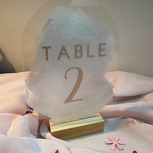 Peut inclure: Panneau de numéro de table en acrylique transparent avec un motif peint en blanc et gris. Le panneau indique "TABLE 2" en couleur or. Le panneau est monté sur une base en bois. Des fleurs décoratives sont dispersées autour du panneau.