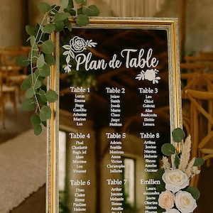 Peut inclure: Un miroir encadré d'or avec une inscription blanche qui dit "Plan de Table" et liste les noms pour chaque numéro de table. Le miroir est sur un chevalet en or avec de la verdure et des fleurs devant.