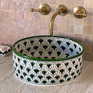 Può includere: Un lavabo da bagno in ceramica bianca con un motivo geometrico verde e nero. Il lavabo ha una forma rotonda e un bordo largo. Il lavabo è montato su una parete e ha un rubinetto in ottone.