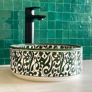Op de afbeelding: Een witte keramische wastafel met een groen en zwart bloemenontwerp. De wastafel is rond en heeft een brede rand. Hij is geplaatst tegen een groene betegelde muur.