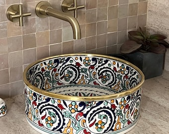 May include: A decorative, round ceramic sink with a colorful floral pattern in blue, red, green, and yellow. The sink has a gold rim and sits on a neutral countertop. A gold faucet and fixtures are mounted on the tiled wall behind it.