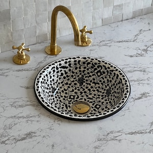 May include: A white ceramic bathroom sink with a black and white floral pattern. The sink has a gold drain and is set on a white marble countertop.