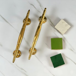 May include: Two gold-toned door handles with a pointed design. The handles are on a white marble surface.