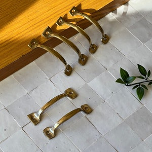 Könnte beinhalten: Eine Sammlung von sechs messingfarbenen Schrankgriffen. Die Griffe haben ein gebogenes Griffdesign mit rechteckigen Montageplatten. Die Hardware ist auf einer weißen Fliesenoberfläche mit Holzkante und einem Zweig grüner Blätter angeordnet.