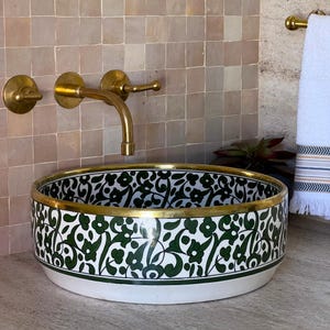 May include: A white ceramic sink with a green and black floral pattern and a gold rim. The sink is round and has a slight curve to the rim. The sink is set against a tiled wall with a gold faucet.
