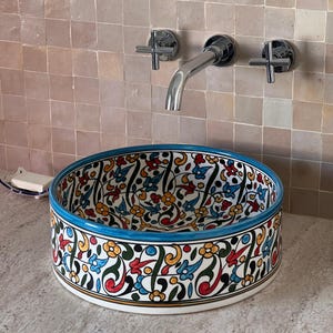 May include: A white ceramic sink with a blue rim and intricate floral patterns in red, yellow, green, and blue. The sink is mounted on a wall with a chrome faucet.