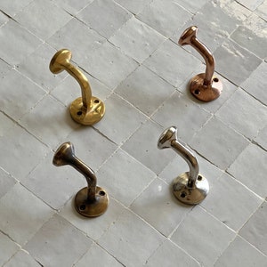 Puede incluir: Cuatro ganchos de pared decorativos en diferentes acabados: oro, cobre, plata y latón. Los ganchos están montados en una pared de azulejos blancos.