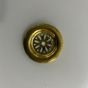 Puede incluir: Una tapa de desagüe de metal de color dorado con un corte decorativo en forma de flor en el centro.