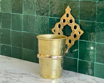 May include: A gold-toned metal toothbrush holder with an ornate design. The holder is mounted to a green tiled wall and has a matching gold-toned cup.