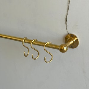 Puede incluir: Un estante para utensilios de cocina de metal dorado con tres ganchos. El estante está montado en una pared y tiene una tapa redonda decorativa.