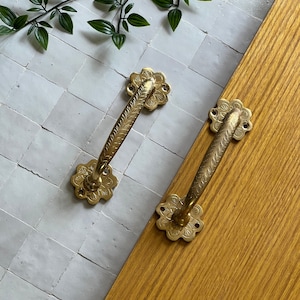 Puede incluir: Dos tiradores de puerta de latón adornados con un diseño floral. Los tiradores están sobre una superficie de azulejos blancos y una superficie de madera.
